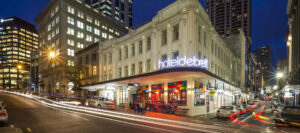 Hotel Debrett, Auckland | Night scene of a busy city street with light trails from moving vehicles, Hotel DeBrett sign illuminated, and people at an outdoor café | New Zealand | The Ultimate Travel Company