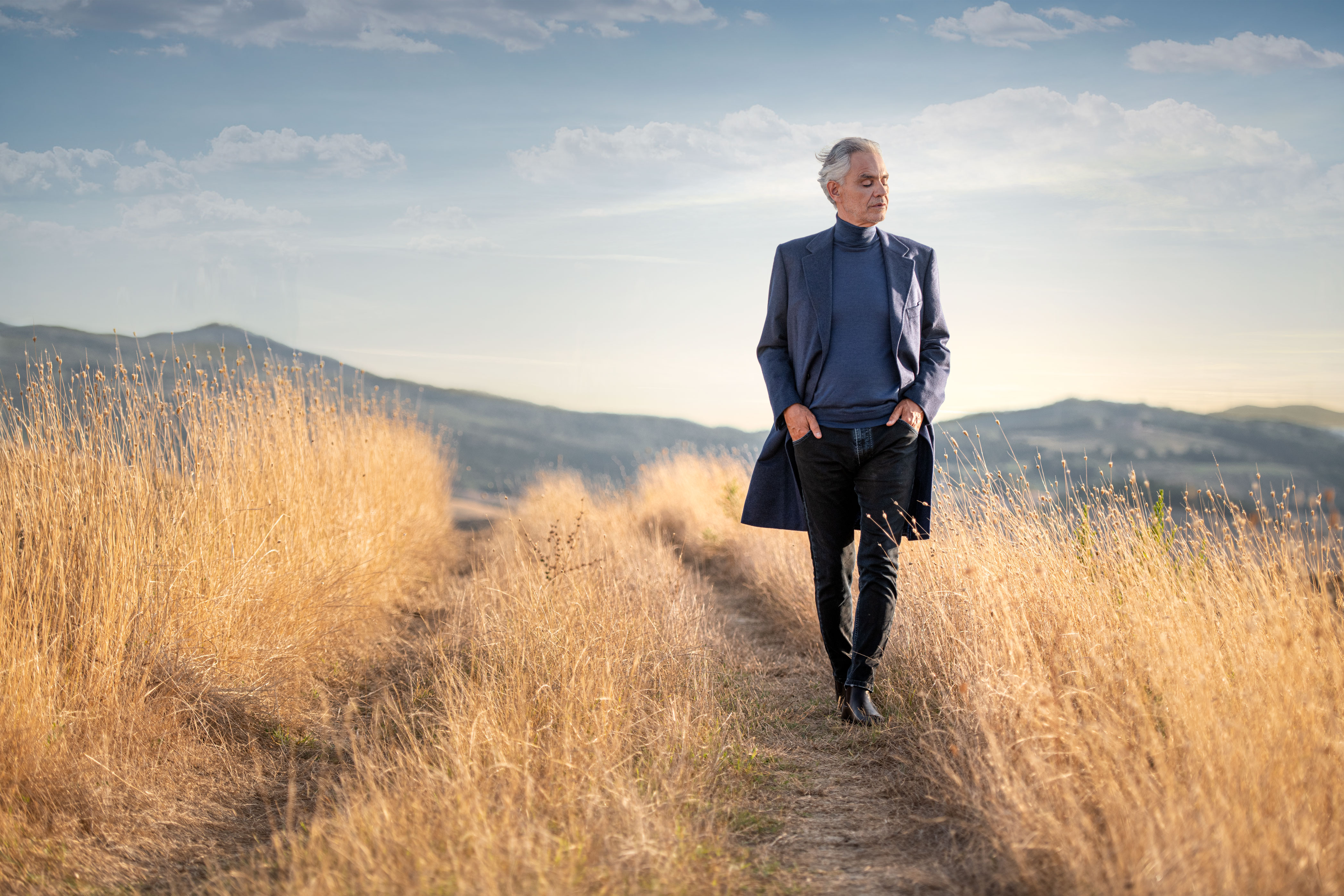 Elegant man walking through a golden field in Tuscany at sunset, wearing a blue coat and turtleneck, with rolling hills and soft sky in the background, luxury travel, Italy countryside, autumn landscape, serene moment.