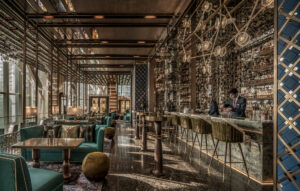 Luxurious interior of Bar Trigona: emerald sofas, marble floors, brass bar stools and mirrored back bar, with two bartenders mixing cocktails in warm natural light.