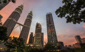 Petronas Twin Towers with skybridge and the Four Seasons Hotel Kuala Lumpur rising over the city at sunset, framed by trees and pastel skies.